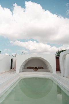 An elegant outdoor area featuring a minimalist architectural style. A small, clear swimming pool takes center stage, surrounded by white walls and arched structures. The sky above is bright with fluffy clouds, adding to the serene ambiance. At the far end, there is a cozy seating area with cushions set in a semi-circular niche.