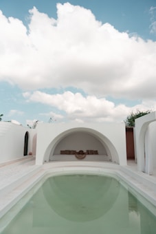 An elegant outdoor area featuring a minimalist architectural style. A small, clear swimming pool takes center stage, surrounded by white walls and arched structures. The sky above is bright with fluffy clouds, adding to the serene ambiance. At the far end, there is a cozy seating area with cushions set in a semi-circular niche.