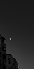 A black and white image featuring a partially visible moon in the sky with several apartment buildings on the left side. The buildings have windows and balconies, with a clear view of architectural details such as railings and window frames.