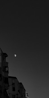 A black and white image featuring a partially visible moon in the sky with several apartment buildings on the left side. The buildings have windows and balconies, with a clear view of architectural details such as railings and window frames.