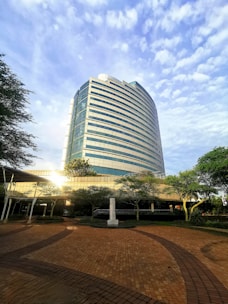 A modern, multi-story glass and steel office building is surrounded by greenery and an open brick walkway. The structure features curved architectural elements and reflective windows that capture the sunlight. Ground-level trees and shrubs provide a touch of nature amidst the urban setting.