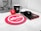 A workspace features a mug with the letter 'H', a red stress ball with 'absa' written on it, a black computer mouse next to a black notebook with a red elastic band and 'absa' logo, and a silver pen. The background includes part of a computer monitor stand and some cables. A red circular mouse pad with the 'absa' logo is prominent on the desk.