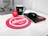 A workspace features a mug with the letter 'H', a red stress ball with 'absa' written on it, a black computer mouse next to a black notebook with a red elastic band and 'absa' logo, and a silver pen. The background includes part of a computer monitor stand and some cables. A red circular mouse pad with the 'absa' logo is prominent on the desk.