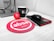 A workspace features a mug with the letter 'H', a red stress ball with 'absa' written on it, a black computer mouse next to a black notebook with a red elastic band and 'absa' logo, and a silver pen. The background includes part of a computer monitor stand and some cables. A red circular mouse pad with the 'absa' logo is prominent on the desk.