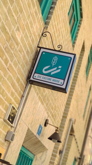 A hanging sign for 'Zee Culture House' is affixed to a yellow-bricked building wall. The sign is predominantly teal with a stylized leaf graphic and white text. The building features teal-framed windows and a decorative light fixture is visible below the sign.