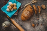 A rustic kitchen scene with a loaf of seeded bread partially sliced on a floured wooden surface. Nearby, a collection of eggs rests on a bright blue cloth, accompanied by a wooden rolling pin, a bottle of milk, wheat stalks, and scattered nuts.