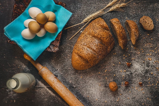 A rustic kitchen scene with a loaf of seeded bread partially sliced on a floured wooden surface. Nearby, a collection of eggs rests on a bright blue cloth, accompanied by a wooden rolling pin, a bottle of milk, wheat stalks, and scattered nuts.
