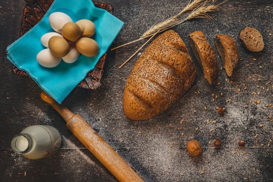 A rustic kitchen scene with a loaf of seeded bread partially sliced on a floured wooden surface. Nearby, a collection of eggs rests on a bright blue cloth, accompanied by a wooden rolling pin, a bottle of milk, wheat stalks, and scattered nuts.
