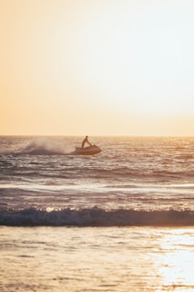 A person rides a jet ski on the ocean, with waves splashing around. The sun is setting in the background, casting a warm golden hue over the water.