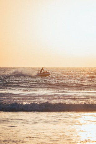 A person rides a jet ski on the ocean, with waves splashing around. The sun is setting in the background, casting a warm golden hue over the water.