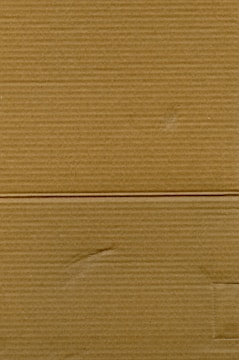 A close-up view of corrugated cardboard with vertical and horizontal lines visible. The texture is coarse, with subtle variations in color and subtle markings suggesting folds or creases.