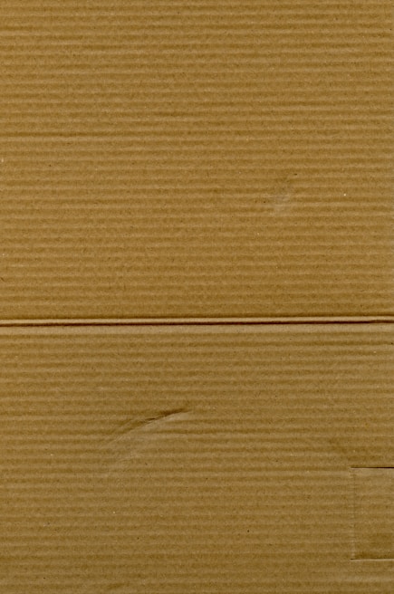 A close-up view of corrugated cardboard with vertical and horizontal lines visible. The texture is coarse, with subtle variations in color and subtle markings suggesting folds or creases.