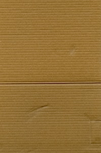 A close-up view of corrugated cardboard with vertical and horizontal lines visible. The texture is coarse, with subtle variations in color and subtle markings suggesting folds or creases.
