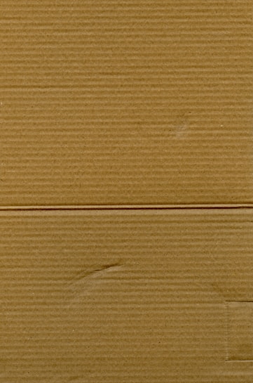 A close-up view of corrugated cardboard with vertical and horizontal lines visible. The texture is coarse, with subtle variations in color and subtle markings suggesting folds or creases.