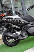 A close-up view of a black V MAX 200 scooter parked inside a showroom. The focus is on the rear wheel and suspension, with the model name 'V MAX 200' clearly visible on the body. The scooter is placed on a green artificial grass mat, and there are reflections in the glass windows behind.