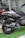 A close-up view of a black V MAX 200 scooter parked inside a showroom. The focus is on the rear wheel and suspension, with the model name 'V MAX 200' clearly visible on the body. The scooter is placed on a green artificial grass mat, and there are reflections in the glass windows behind.