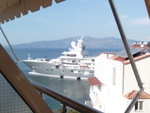 A large luxury yacht is sailing on a calm body of water, viewed from a terrace with awnings and part of a building with white walls and terracotta roofs. The backdrop features distant mountains and a clear blue sky.