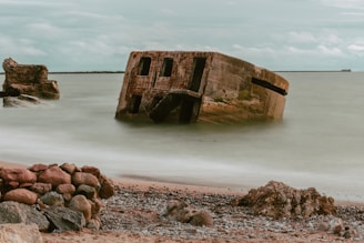 A partially submerged concrete structure rests at an angle in the ocean near a rocky shore. The sky is overcast, casting a muted light over the scene. The sea appears calm, with gentle waves approaching the scattered rocks and pebbles on the beach.