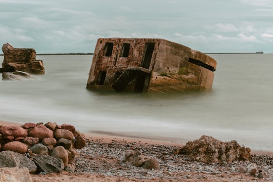 A partially submerged concrete structure rests at an angle in the ocean near a rocky shore. The sky is overcast, casting a muted light over the scene. The sea appears calm, with gentle waves approaching the scattered rocks and pebbles on the beach.