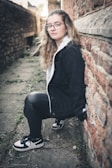 A young person with long wavy hair, wearing glasses, a black jacket with a white inner lining, black leather pants, and black and white sneakers is leaning against a rough, reddish-brown brick wall. The scene appears to be in a narrow alleyway with stone pavement and some greenery sprouting through the cracks.