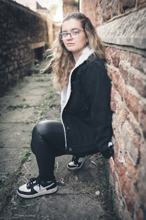 A young person with long wavy hair, wearing glasses, a black jacket with a white inner lining, black leather pants, and black and white sneakers is leaning against a rough, reddish-brown brick wall. The scene appears to be in a narrow alleyway with stone pavement and some greenery sprouting through the cracks.