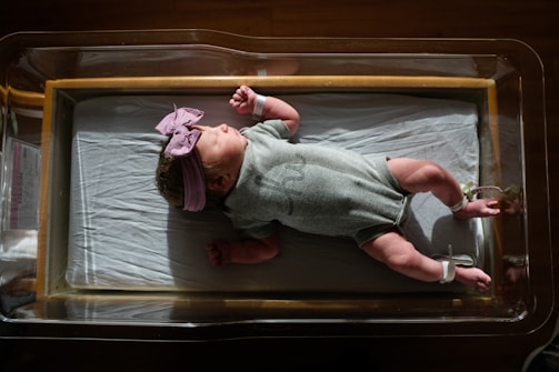 A newborn baby is resting in a hospital bassinet, wearing a gray onesie and a pink bow headband. The light casts soft shadows, highlighting the baby's peaceful expression.