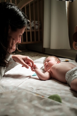 A woman gently holds a baby's hand as the baby lies on a soft play mat. The scene is set in a cozy, softly lit room with a crib in the background. The baby is wearing a diaper and looking up at the woman with curiosity.