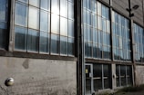 A large industrial building facade with numerous tall glass windows spanning multiple floors. The windows are divided into smaller panes and show signs of wear, with visible streaks and smudges. The concrete wall beneath the windows has patches and a metal grid leaning against it. A door with safety signs is located near the center, and there are plants growing along the base of the building.