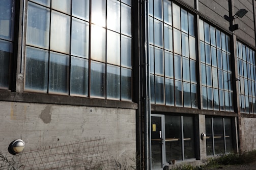 A large industrial building facade with numerous tall glass windows spanning multiple floors. The windows are divided into smaller panes and show signs of wear, with visible streaks and smudges. The concrete wall beneath the windows has patches and a metal grid leaning against it. A door with safety signs is located near the center, and there are plants growing along the base of the building.