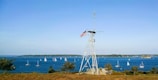 A maritime scene featuring a tall structure with an American flag atop it, positioned on a grassy area. Numerous sailboats with white sails are visible on the blue water. The horizon shows a distant shoreline under a clear blue sky.