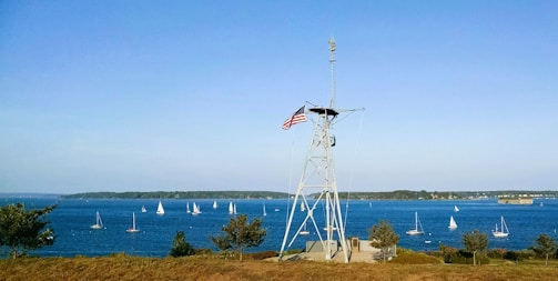 A maritime scene featuring a tall structure with an American flag atop it, positioned on a grassy area. Numerous sailboats with white sails are visible on the blue water. The horizon shows a distant shoreline under a clear blue sky.