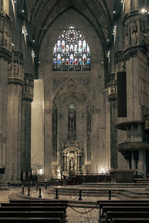 An ornate interior of a cathedral showcasing towering stone columns, intricately designed arches, and a large stained glass window above an elaborate altar. The architecture is grand and majestic, conveying a sense of historical reverence, with dim lighting accentuating the solemn atmosphere.