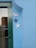 A light blue wall features a green door with a black handle. Above the door are metal address numbers '575'. Two decorative starfish, one blue and one white, are mounted on the wall next to the door. A reflection in the door's glass panel shows part of a neighboring building with a tiled roof.