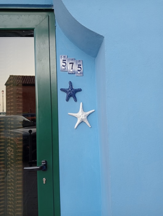 A light blue wall features a green door with a black handle. Above the door are metal address numbers '575'. Two decorative starfish, one blue and one white, are mounted on the wall next to the door. A reflection in the door's glass panel shows part of a neighboring building with a tiled roof.