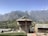 A rustic building with stone architecture, set against a backdrop of majestic mountains. The roof is equipped with a solar panel, and the surrounding landscape is dotted with greenery and other small structures. The scene is tranquil and bathed in sunlight, with a clear blue sky.