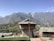 A rustic building with stone architecture, set against a backdrop of majestic mountains. The roof is equipped with a solar panel, and the surrounding landscape is dotted with greenery and other small structures. The scene is tranquil and bathed in sunlight, with a clear blue sky.