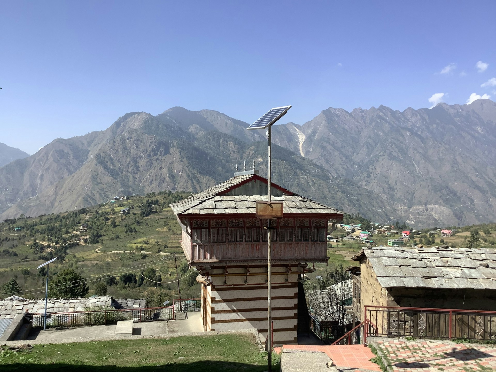A rustic building with stone architecture, set against a backdrop of majestic mountains. The roof is equipped with a solar panel, and the surrounding landscape is dotted with greenery and other small structures. The scene is tranquil and bathed in sunlight, with a clear blue sky.