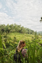 A person with a backpack stands amidst lush green rice terraces, holding a smartphone. Palm trees line the background, and the sky is partly cloudy, contributing to a serene landscape.