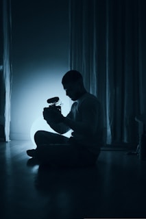 A person is sitting cross-legged on the floor in a dimly lit room, holding a camera with a microphone attached. The silhouette is illuminated by a soft blue light, creating a serene atmosphere with curtains in the background.