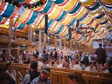 A bustling festival tent with colorful drapes in red, yellow, green, and blue adorning the ceiling. The scene is lively with many people gathered at long wooden tables, engaging in conversation and enjoying the festive atmosphere. A traditional band performs on a stage, adding to the vibrant mood. Traditional decorations and lights enhance the ambiance.