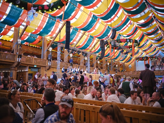 A bustling festival tent with colorful drapes in red, yellow, green, and blue adorning the ceiling. The scene is lively with many people gathered at long wooden tables, engaging in conversation and enjoying the festive atmosphere. A traditional band performs on a stage, adding to the vibrant mood. Traditional decorations and lights enhance the ambiance.