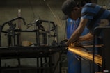 A person wearing a cap and striped shirt is using a power tool to grind or cut metal, producing bright orange sparks in a dimly lit workshop. Metal frames and blue barrels are visible in the background.