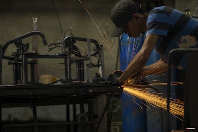 A person wearing a cap and striped shirt is using a power tool to grind or cut metal, producing bright orange sparks in a dimly lit workshop. Metal frames and blue barrels are visible in the background.
