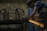 A person wearing a cap and striped shirt is using a power tool to grind or cut metal, producing bright orange sparks in a dimly lit workshop. Metal frames and blue barrels are visible in the background.