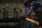 A person wearing a cap and striped shirt is using a power tool to grind or cut metal, producing bright orange sparks in a dimly lit workshop. Metal frames and blue barrels are visible in the background.