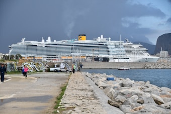 A large cruise ship is docked at a port with a cloudy sky overhead. People are walking along a pathway beside the water, and there is a section with graffiti on the left. The scene includes a stone-lined shoreline and distant mountains in the background.