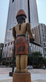 A large wooden sculpture of a figure stands in an urban environment, facing a tall modern building. The sculpture is intricately designed with patterns on its clothing and appears to have a traditional or cultural style. The surrounding area includes other buildings, trees, and a street with a car.