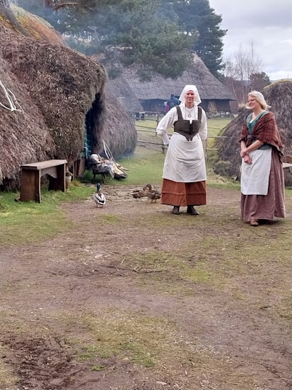 Two individuals dressed in historical clothing stand in front of thatched-roof huts, suggesting a setting from an earlier time period. Ducks can be seen walking along the dirt path, and a bench is positioned near the entrance of one of the huts. Trees and other huts can be seen in the background under a cloudy sky.