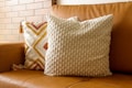 Two decorative pillows are placed on a brown leather sofa. One pillow features a textured, knitted design in cream, while the other has a geometric pattern with orange and yellow thread detailing. The background consists of a brick wall in a light brown hue.