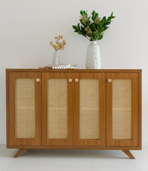 A wooden sideboard with woven rattan panels and modern legs is adorned with a large white vase containing dense green foliage. A smaller white vase with delicate orange flowers and a decorative string of white beads is also present.